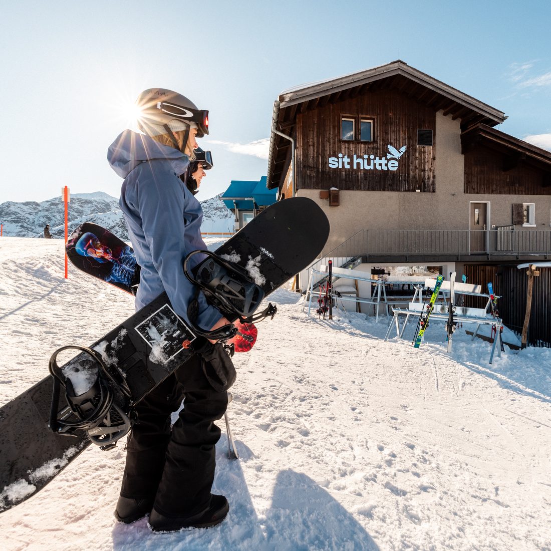 Zwei Snowboarderinnen in der Sonne vor der sit hütte mit Schriftzug, dahinter die Berge